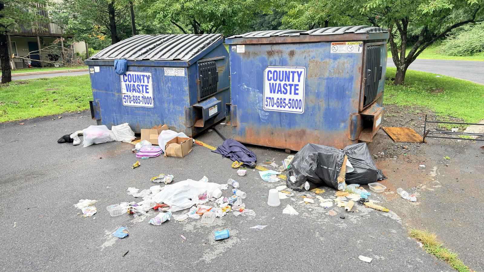 Bagged and loose rubbish scattered across the asphalt around two overflowing dumpsters